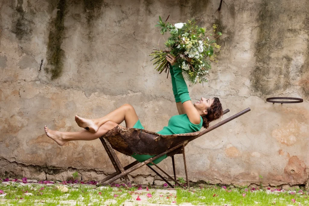 Kobieta leżąca w taczce unosi bukiet kwiatów – kreatywna i artystyczna interpretacja florystyki