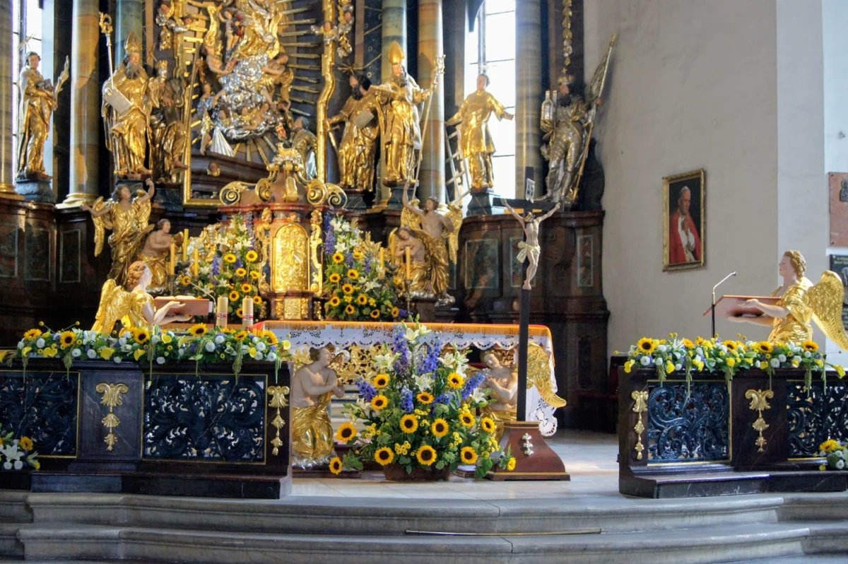 Dekoracja kwiatowa ołtarza w barokowym kościele z kompozycjami ze słoneczników, lilii i kwiatów sezonowych otaczających prezbiterium.
