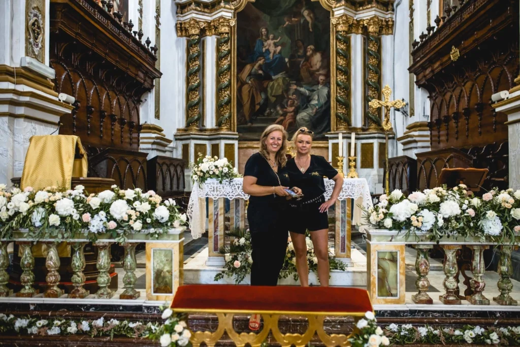 Florystki prezentujące dekorację kwiatową ołtarza w zabytkowym kościele z kompozycjami z białych kwiatów i zieleni.