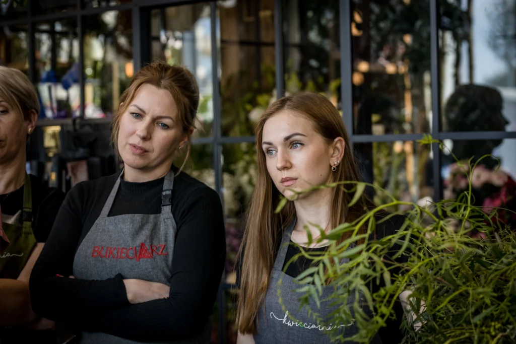 Florystki z zespołu Bukieciarz w fartuchach firmowych, stojące w kwiaciarni w otoczeniu zielonych roślin.
