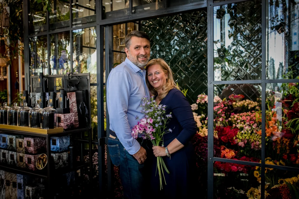 Andrzej i Monika Dąbrowscy w otoczeniu kwiatów, Monika trzyma bukiet fioletowych i różowych kwiatów.